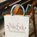 Stofftasche aus Leinen mit Logo „NaSch NaturKosmetik“ auf einem Stuhl.